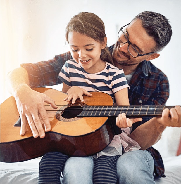 Young girl sitting in her dad’s lap while they play guitar.